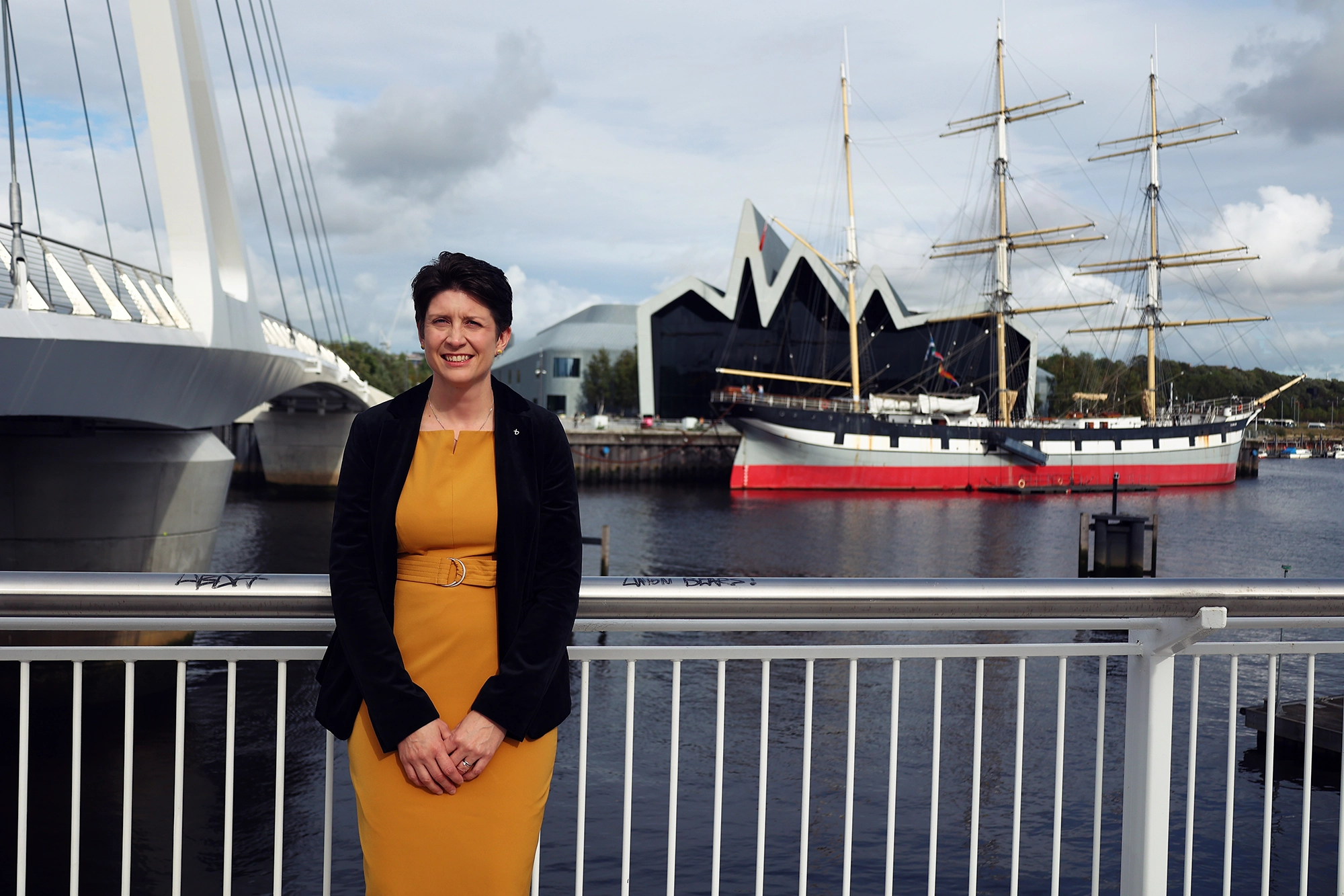 Alison Thewliss standing on the riverside in Govan, with the Tall Ship and Riverside Museum in the background.